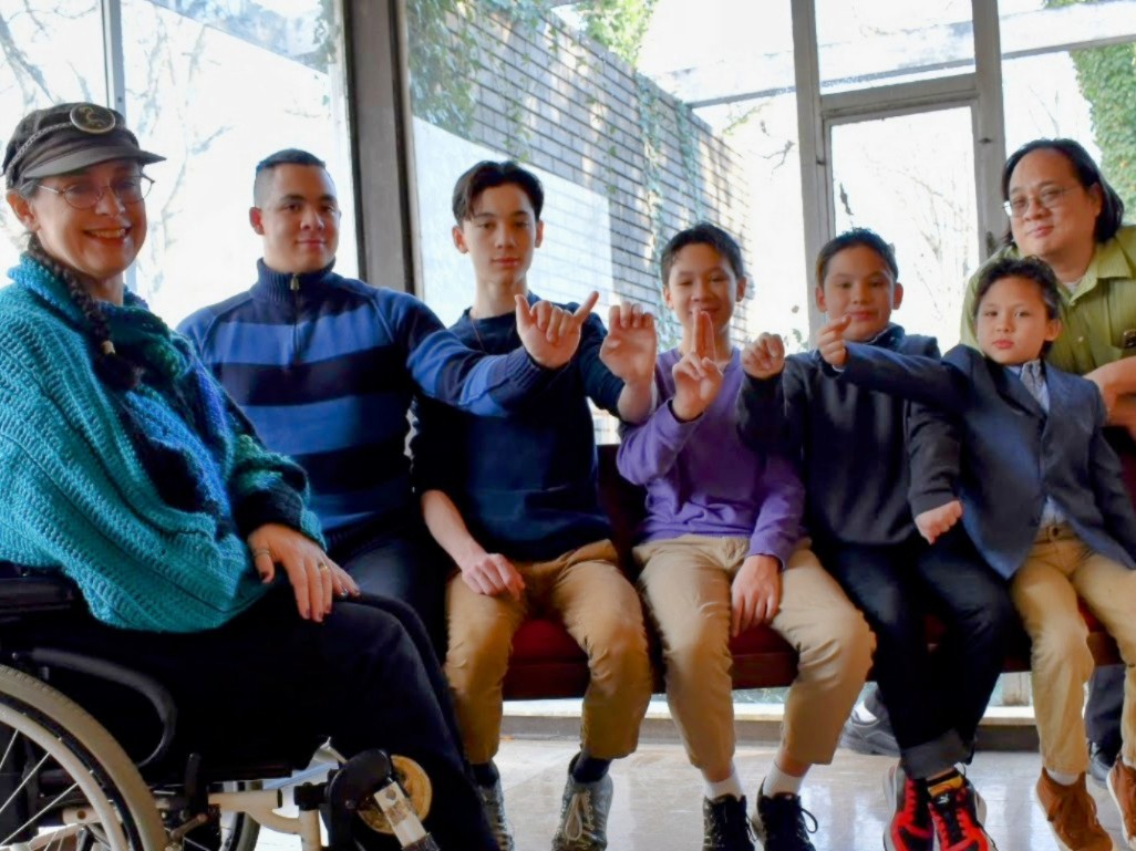 an image of Ahmie in her wheelchair with her sons William, Delano, Lincoln, Ulysses, and Quincy fingerspelling YEUNG and their father Garvin leaning in from the right edge of the frame